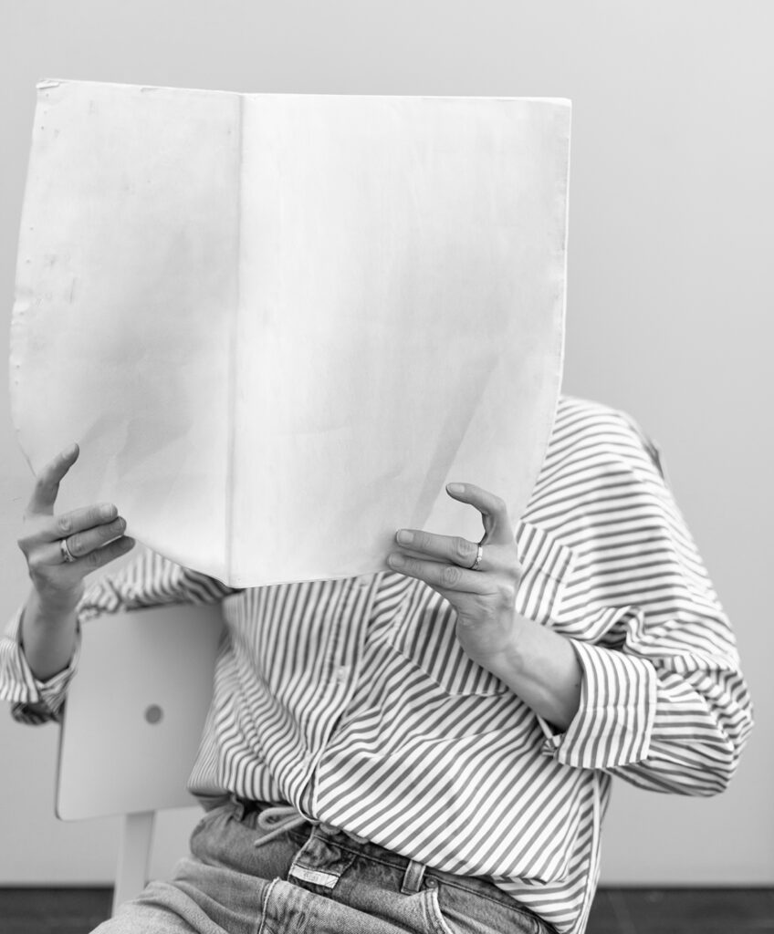 About Blank, keine Angst vor dem weissen Blatt, Frau lehnt auf Stuhl und hält eine Zeitung vor das Gesicht mit leeren Seiten